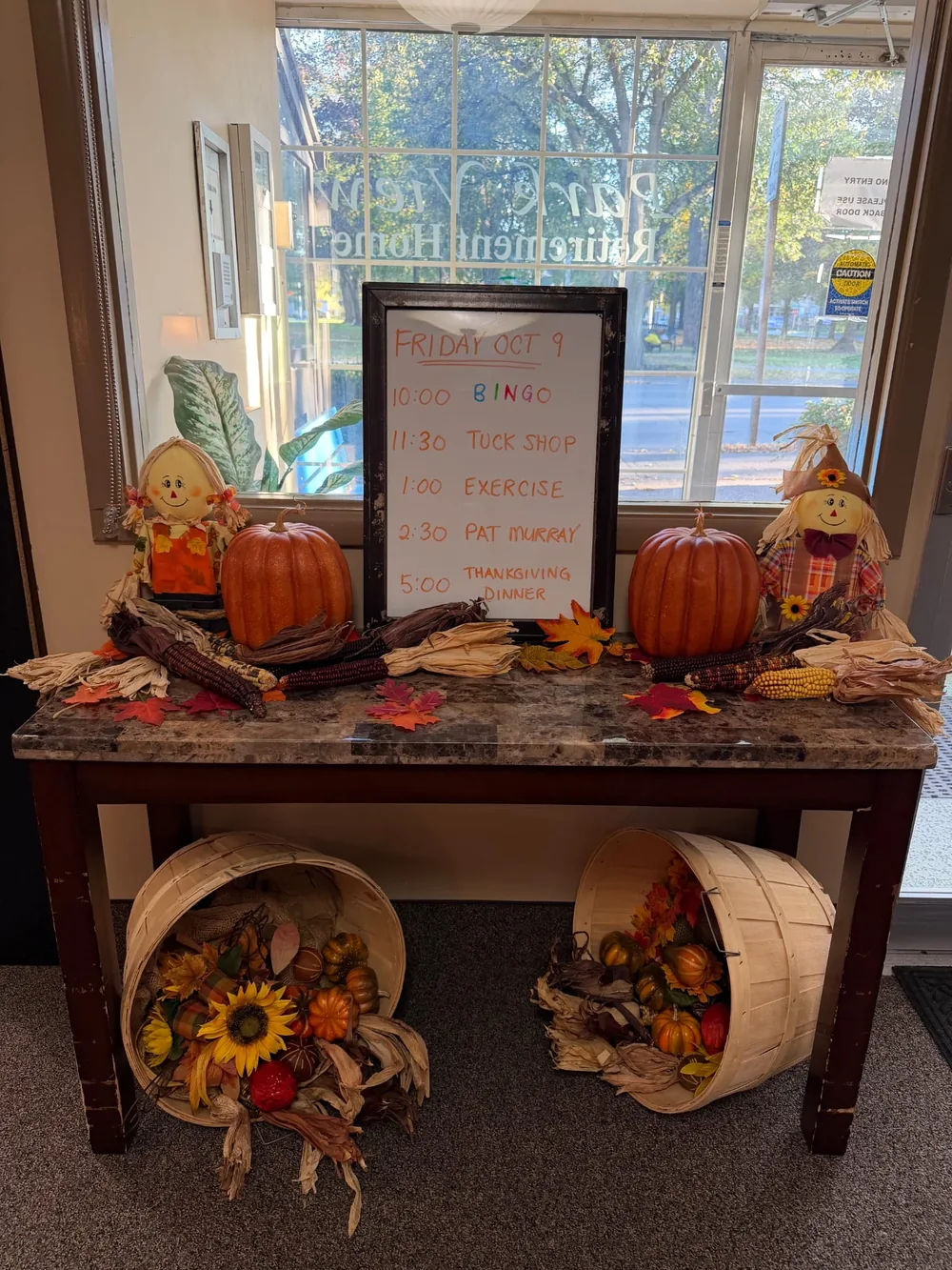 Lobby table decorated with pumpkins, scarecrows and corn beside a whiteboard listing the day's activities and Thanksgiving dinner