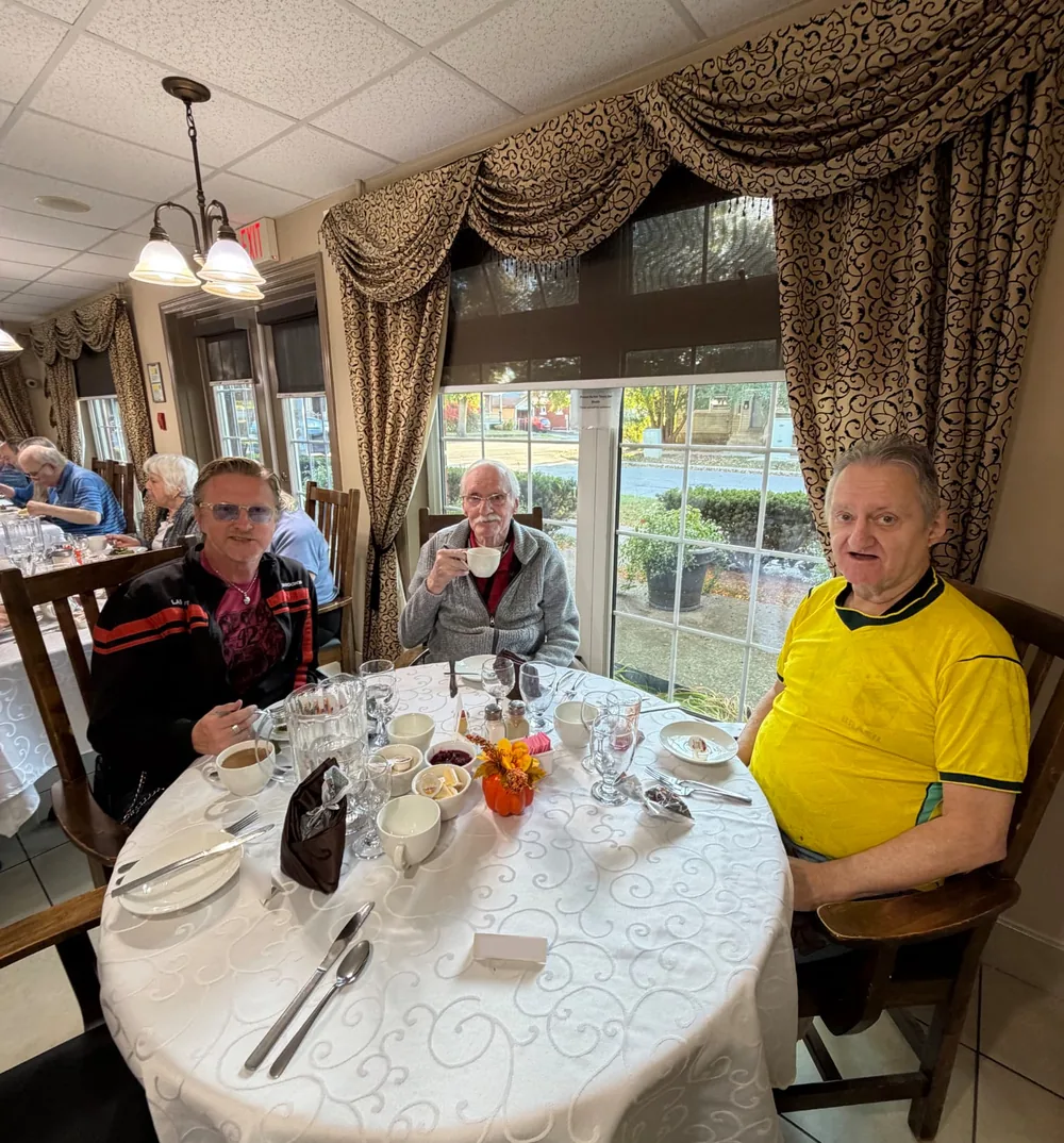 Three men at a round table by the window, one raising a coffee cup, during Thanksgiving dinner
